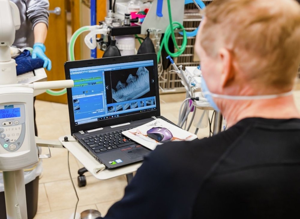 A person wearing a mask looks at a laptop displaying dental x-rays, with medical equipment in the background.