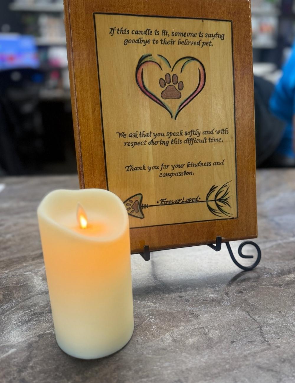 A lit candle sits next to a wooden plaque with a poem about saying goodbye to a pet. The plaque features a heart with a paw print inside.